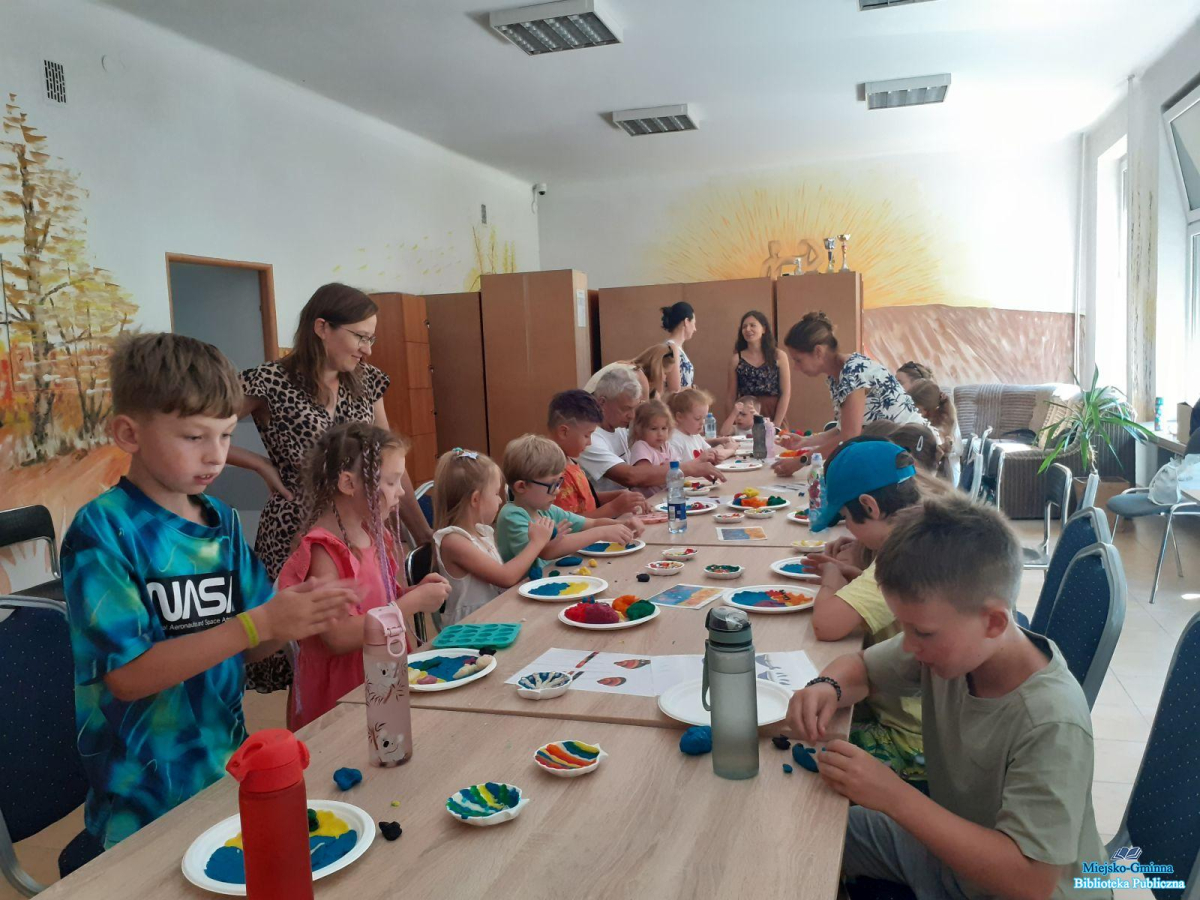 Grupa dzieci siedzi przy stoliku a dwójka z nich na stojąco wygniata ciastolinę i dekorują gipsowe muszelki Grupa dzieci siedzi przy stoliku a dwójka z nich na stojąco wygniata ciastolinę i dekorują gipsowe muszelki