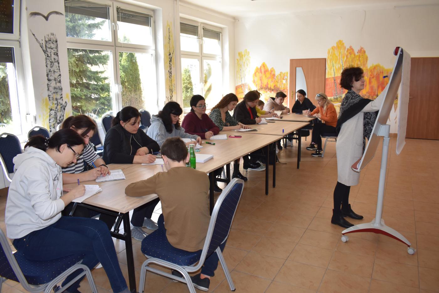 12 osób siedzi przy stoliku a Pani pisze na tablicy