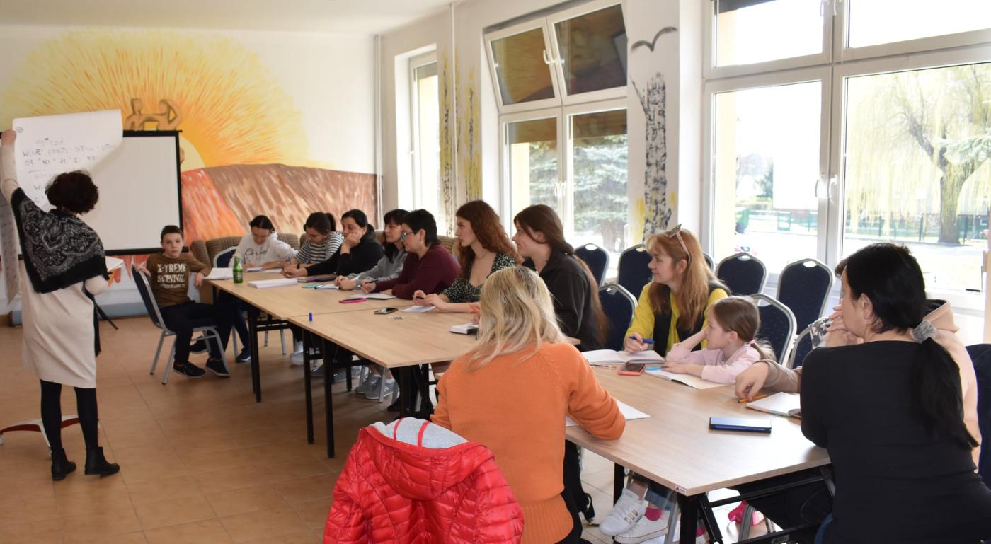 13 osób siedzi przy stoliku a jedna Pani stoi przy tablicy