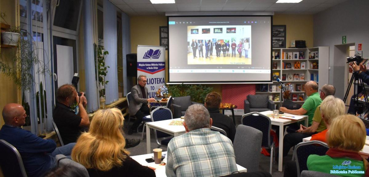 Pan siedzi i omawia zdjęcie na slajdzie a zebrani goście siedzą przy stolikach