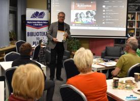 Pan stoi i trzyma w dłoniach książkę a zebrani goście siedzą przy stolikach