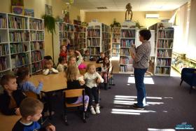 Pani mówi a dzieci siedzą przy stoliku i dziewczynek trzyma rękę w górze