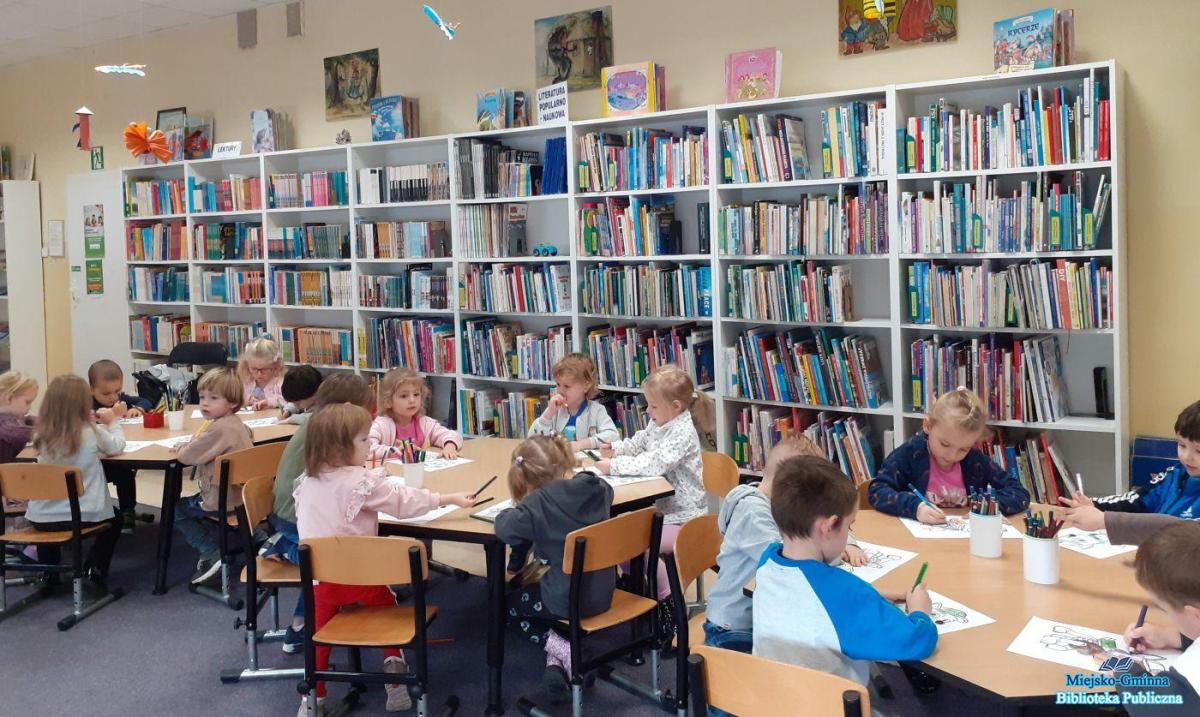 Dzieci siedzą przy trzech stoliczkach i rysują w tle widać regały z książkami