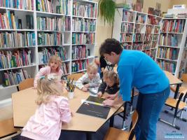 Pani w niebieskim swetrze stoi przy stoliku przy którym siedzą cztery dziewczynki