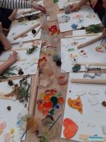 Na stolkiu leżą prace plastyczne oraz farbki i kubeczki z wodą