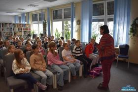 Pani stoi i powiada dzieciom, które siedzą na krzesłach