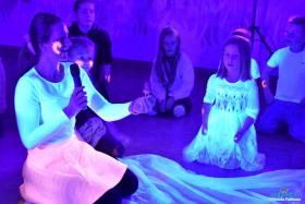 Pani siedzi an podłodze  w ręce trzyma mikrofon i małą figurkę a obok siedzi dziewczynka