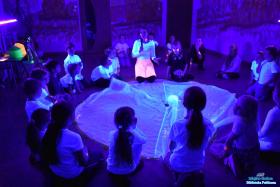 Pani z dziećmi siedzą na podłodze a przed nimi leży duża chusta