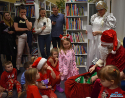Dzieci siedzą na podłodze, rodzice i Pani w białej sukience stoi, a Mikołąj siedzi w fotelu