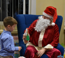 Mikołaj siedzi w fotelu i daje małemu chłopcu w niebieskiej koszuli prezent