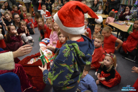 Chłopiec w czapce Mikołąja trzyma w dłoniach zielone pudełko, a w tle widać inne dzieci