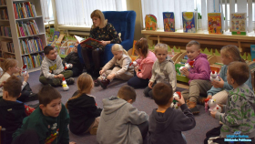 Dzieci siedzą na dywanie, a Pani siedzi na fotelu i czyta książkę