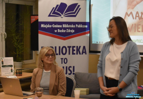 Dwie Panie przy stoliku, jedna z nich stoi, druga siedzi na krześle