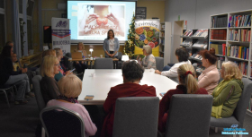 Pani stoi a uczestnicy spotkania siedzą przy stole