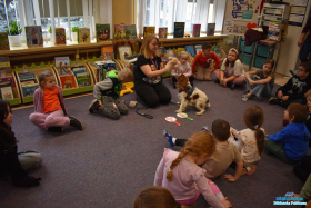 Pani ma na dłoni pacynkę, a pies siedzi przed nią. Wokół siedzą dzieci