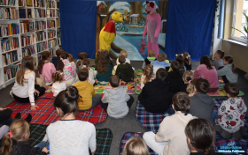 Dwaj aktorzy stoją, a dzieci siedzą na dywanie