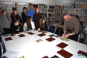 Pan otwiera książkę, która leży na stoliku, a młodzież stoi obok