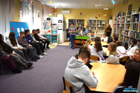 Młodzież siedzi przy stolikach a Pani stoi i mówi do nich