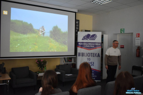 Pan stoi i mówi a w tle wyświetlony jest fragment filmu