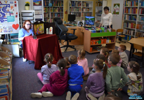 Jedna Pani czyta, druga stoi obok półek, a dzieci siedzą na podłodze