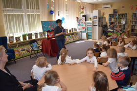 PANI STOI I TRZYMA KSIĄŻKĘ A DZIECI SIEDZĄ