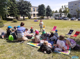 Pani stoi i czyta dzieciom, które siedzą na kocach na trawie