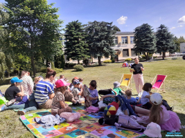Pani w czarnej bluzce czyta dzieciom, któe siedzą na trawie przed budynkim biblioteki