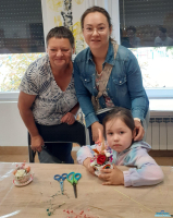 Dwie dorosłe kobiety stoją, a dziewczynka siedzi przy stole trzymając filiżankę