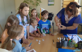 Dzieci stoją przy biurku a Pani wyjmuje z pojemniczka karteczkę