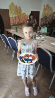 Dziewczynka stoi przy stoliku i prezentuje wykonane prace z kolorowej ciastoliny, dwoje dzieci siedzi przy stoliku a obok stoi osoba dorosła