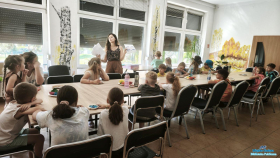 Grupa dzieci siedzi przy stolikach  z  osobą dorosłą, dwoje dzieci stoi przy stoliku przy oknie a pani  pokazuje obrazkowe instrukcje potrzebne do wykonywania ćwiczeń z ciastoliną