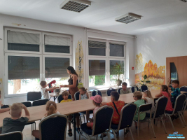 Grupa dzieci siedzi przy stolikach a obok stoi pani z dzieckiem  przy blenderze leżącym na stoliku obok przy oknie i mieszają składniki do masy, z której będzie ciastolina