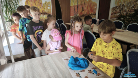 Grupa dzieci stoi przy stoliku w kolejce do wyrabiania niebieskiej ciastoliny a jeden chłopiec trzyma w rękach ciastolinę i formuje z niej elementy do podwodnego świata