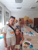 Mama z córeczką stoją przy stoliku, na którym znajdują się prace dziewczynki - muszla wypełniona ciastoliną i podwodny świat z ciastoliny na papierowym talerzyku, a w tle uczestnicy zajęć