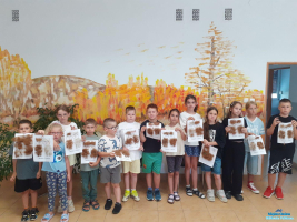 Grupa dzieci stoi w pomieszczeniu biblioteki i trzyma w rękach swoje prace plastyczne