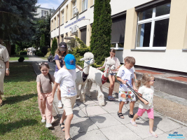 Grupa dzieci prowadzi dwie alpaki na smyczy przy budynku biblioteki