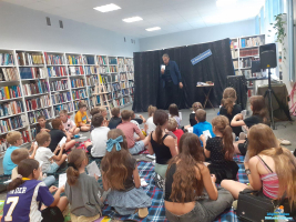Pan stoi i trzyma kartkę, a dzieci siedzą