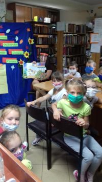 Pani Marta prezentuje książkę dzieciom, które siedzą przy stoliku w bibliotece