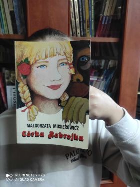 Dziewczynka pozuje do zdjęcia z książka na tle polek z książkami